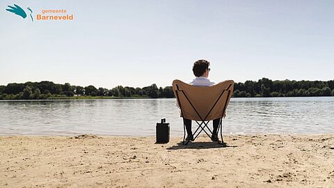 Videostill van burgemeester Jacco van der Tak op een stoel aan recreatieplas Zeumeren. Logo van gemeente Barneveld in de linker bovenhoek.