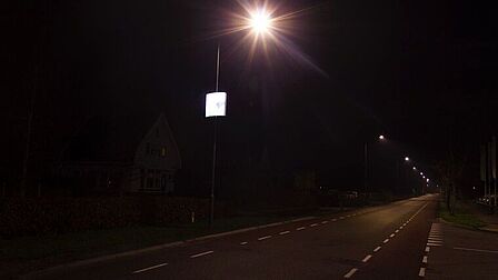 Terschuur, weg in de avond met lantaarnpalen. Aan 1 lantaarnpaal hangt een bord met verlichte reclame. Daar richt je oog zich op